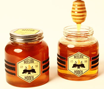 Two jars of honey with metal lid and a honey spoon with white labels.