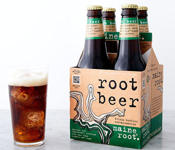 A four-pack of bottles in a beige box and a glass filled with root beer.